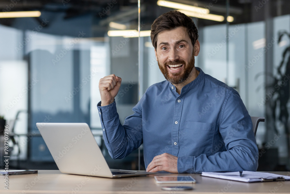 Fototapeta premium Portrait of a happy young man rejoicing in achievement and success showing a yes gesture with his hand. Sitting at the desk in the office and looking at the camera