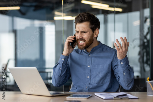 Angry and irritated young businessman sitting in the office at the desk and talking on the phone while waving his hands photo