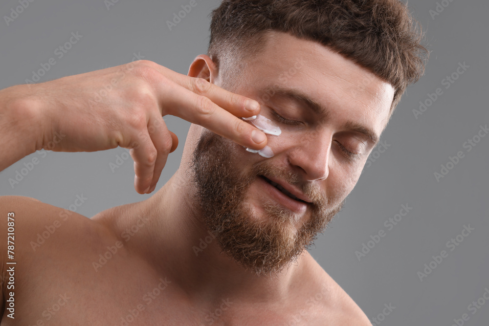 Obraz premium Handsome man applying moisturizing cream onto his face on grey background