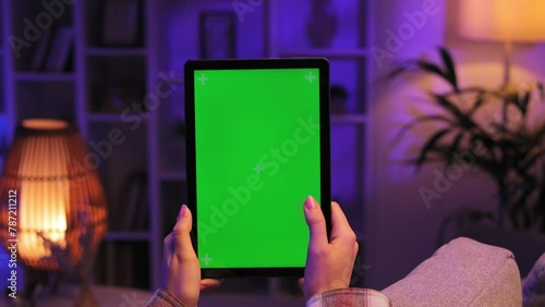 Caucasian girl scrolls sideways and taps the green screen of a tablet with her thumb, lying on the sofa in the evaning in a cozy living room. Close up of a female hand holding a mobile phone. photo