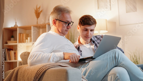 Network search. Family support. Internet technology. Senior father learning to use laptop program together with son home room interior.