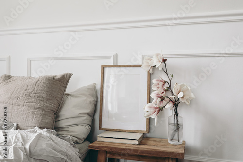 Empty vertical wooden picture frame mockup, old books. Wooden night stand with fluted glass vase. Blooming magnolia tree branches. Scandinavian interior, bedroom. White wall background, side view © tabitazn