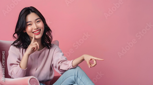 Young elegant Asian woman smiling and pointing to empty copy space and sitting on armchair isolated on pink background : Generative AI © Generative AI