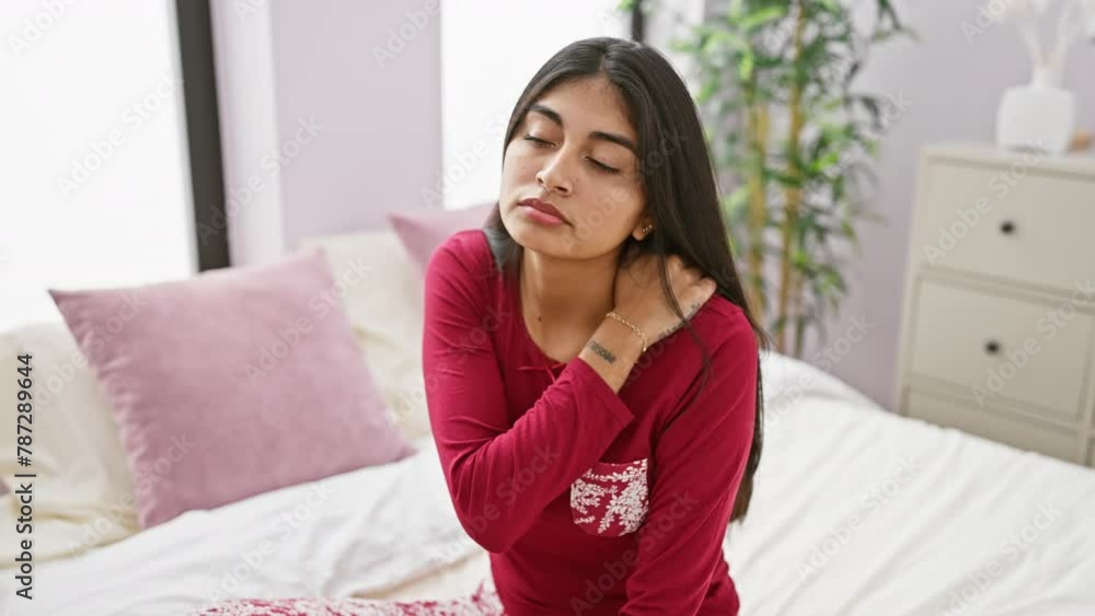 A young south asian woman in red pajamas stretching while sitting on a bed in a bright bedroom. A young south asian woman in red pajamas stretching while sitting on a bed in a bright bedroom.