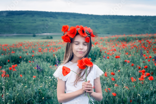 Portrait of a beautiful little girl with a flower wreath on her head on the background of a blossomed poppy field before the sunset.