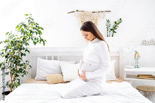 A pregnant woman in a white dress with a big belly strokes him with her hand. The expectant mother is waiting and preparing for the birth of a child at home on a bed in a bright bedroom