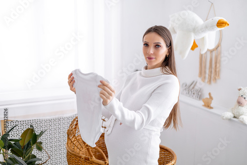 A pregnant woman with a big belly looks at baby clothes for a newborn. The expectant mother is waiting and preparing for the birth of a child at home in a bright children's bedroom by the crib