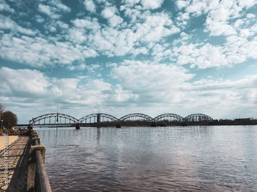 Cityscape image of a beautiful bridge over a calm Daugava river under a clear blue sky with fluffy white clouds, showcasing stunning architectural design and tranquil natural surroundings of Riga, Lat photo