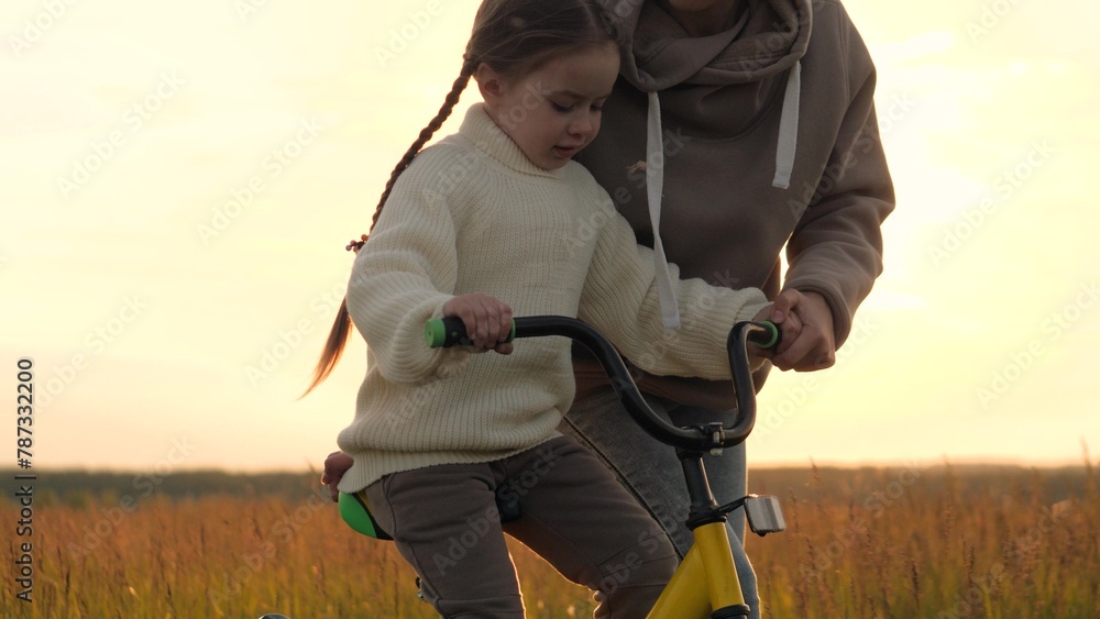 Woman teaching girl to ride bike, silhouettes at sunrise in field. Emotional support, participation, presence and encouragement from mother in learning process to help child overcome fear of failure. - obrazy, fototapety, plakaty 