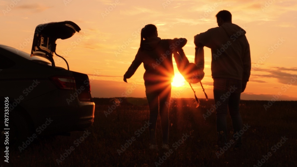 Silhouettes of having fun mom dad kid. Golden sunset, cheerful joyful family at holidays holding hands, lifting baby girl high in air, playing together. Fun active game. Traveling by car to nature. - obrazy, fototapety, plakaty 