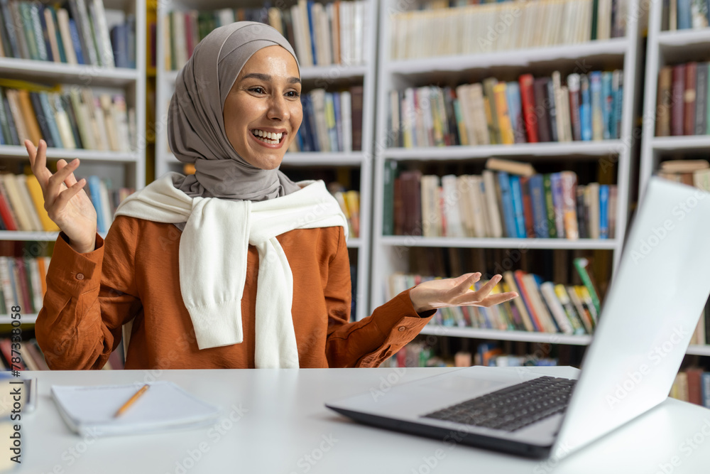 A vivacious young woman wearing a hijab is smiling while engaging in an animated video call. She sits in a library, surrounded by books, exuding a friendly demeanor. A vivacious young woman wearing a hijab is smiling while engaging in an animated video call. She sits in a library, surrounded by books, exuding a friendly demeanor.