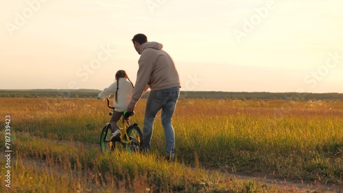 Parent teaches child to ride bicycle. Father and daughter in field at epic sunset. Child safety, dad regulates and controls beloved girl kid on bike, warning against injuries and bruises. Slow motion © Валерий Зотьев