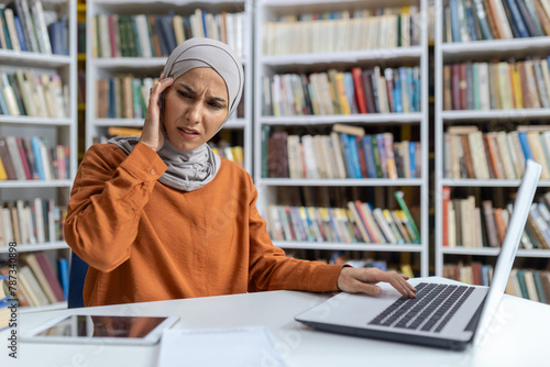 Worried young woman wearing hijab feeling stressed while studying in a library, surrounded by books. She is using a laptop and has a troubled expression.