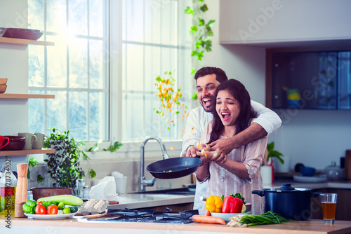 Obraz na plátně Asian Indian happy couple having fun while tossing vegetable in pan in kitchen