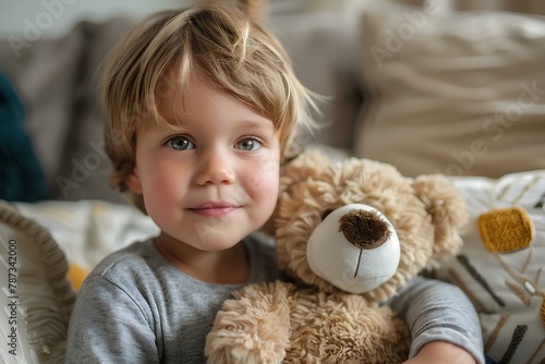 A little boy holding a teddy bear on a bed with pillows and pillows behind him smiling at the
