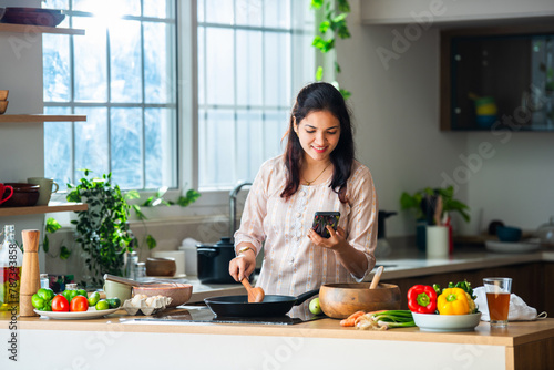 Indian asian woman cooking and using smartphone in kitchen