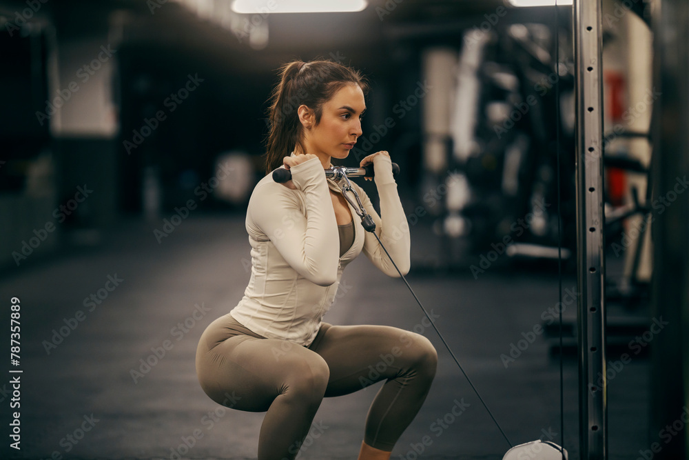 A fit athlete is practicing her upper body with cable machine in squatting position in a gym.