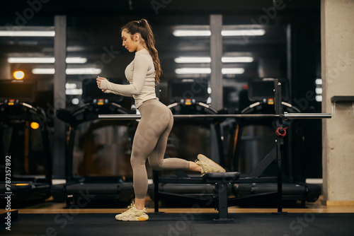 A fit sportswoman is doing legs dips on a bench in a gym.