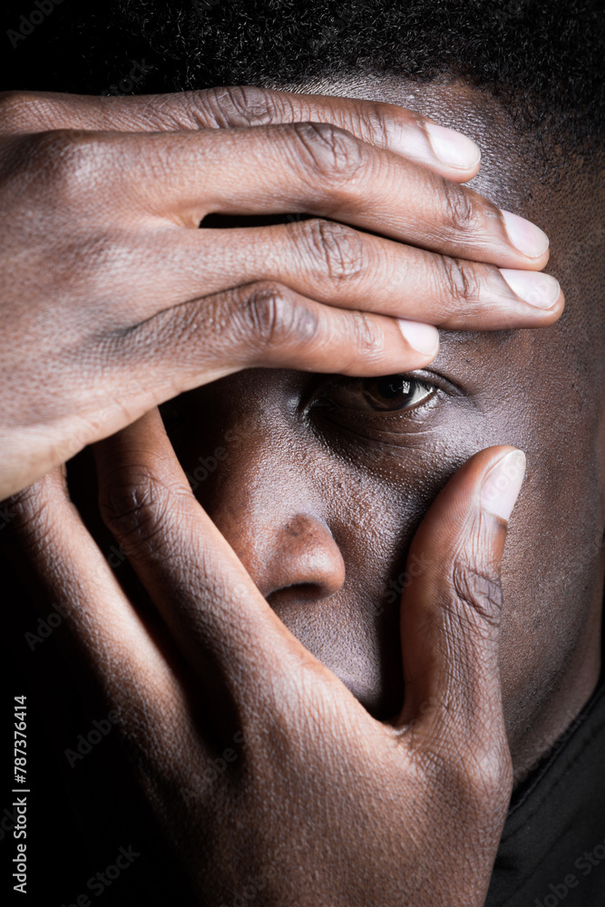 Fototapeta premium A black man covers his face with his hand, revealing one eye and expressing contemplation.