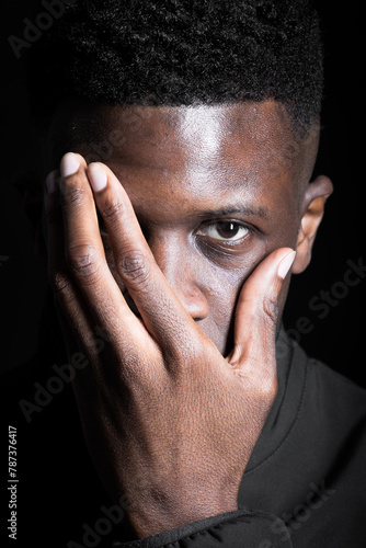 A man covers half of his face with his hand, eyes gazing intently into the camera against a black backdrop.