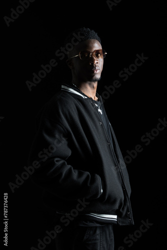 A black man gazes calmly to the side against a dark backdrop, clad in stylish attire.