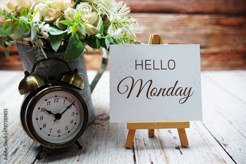 Hello Monday text message written on paper card with wooden easel and alarm clock with flower in metal vase decoration