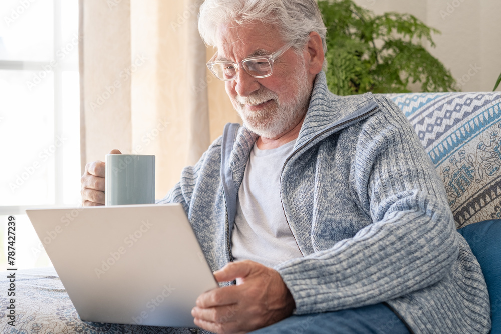 Senior bearded smiling man sitting on sofa looking at laptop holding a coffee cup - indoor, at home concept - caucasian elderly retired man using technology Senior bearded smiling man sitting on sofa looking at laptop holding a coffee cup - indoor, at home concept - caucasian elderly retired man using technology