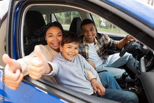 Happy family of three, mother, son and father driving car, woman and boy showing thumbs up in open window, enjoying time together at weekend