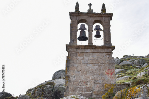 A stone bell tower with two bells in separate arches, situated on rocky terrain with moss and lichen. The tower has a cross at the top and red markings on the lower right side. The sky is overcast. photo