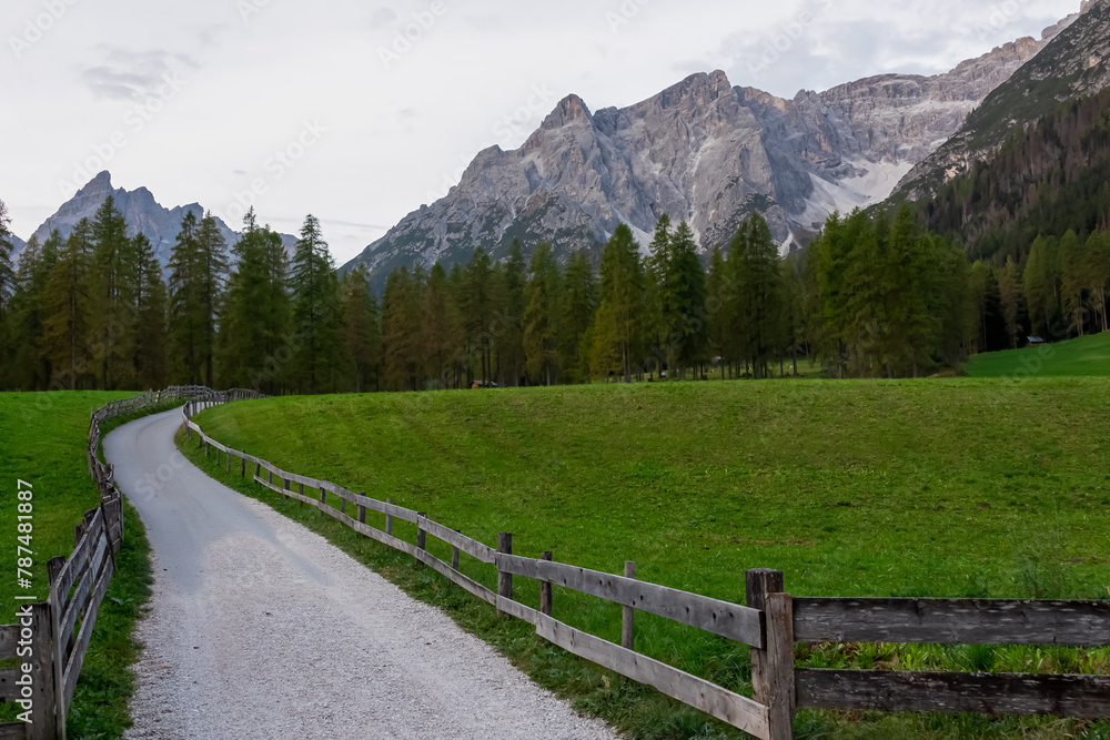Scenic hiking trail along alpine meadow secured by wooden fence in panoramic valley Fischleintal near Moos. Panoramic view of majestic mountain ridges of Sexten Dolomites in Italian Alps. Wanderlust