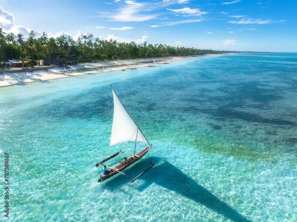 Obraz premium Aerial view of the sailboat on blue sea, empty white sandy beach at sunset. Summer vacation in Zanzibar. Tropical landscape with boat, ocean with clear water, green palms, sky. Top drone view. Exotic