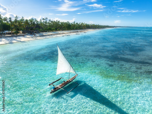 Aerial view of the sailboat on blue sea, empty white sandy beach at sunset. Summer vacation in Zanzibar. Tropical landscape with boat, ocean with clear water, green palms, sky. Top drone view. Exotic