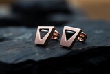 Two modern geometric stud earrings in rose gold are placed on top of a rugged rock surface. The gold rings shimmer under the natural light, contrasting with the rough texture of the rock