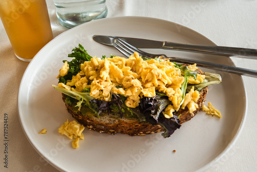 avocado toast with fresh greens and scrambled eggs on top of whole grain bread on a white plate with fork and knife. Healthy breakfast concept