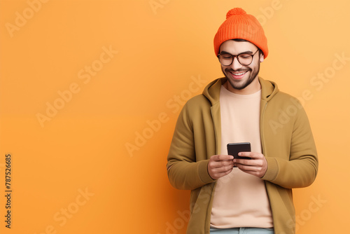 A young man in a trendy orange beanie and glasses smiles as he uses his smartphone, dressed in casual autumn attire against a vibrant orange background.