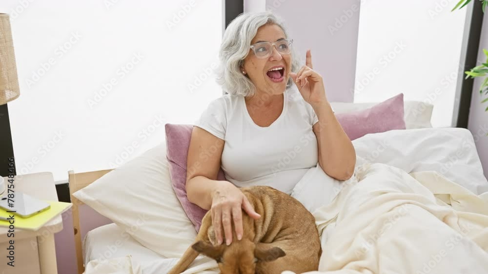 Surprised middle age grey-haired woman, delighted by a brilliant idea! lounging in pyjamas on bed with dog, her finger pointing in excited eureka moment, displaying her happy face in bedroom. Surprised middle age grey-haired woman, delighted by a brilliant idea! lounging in pyjamas on bed with dog, her finger pointing in excited eureka moment, displaying her happy face in bedroom.