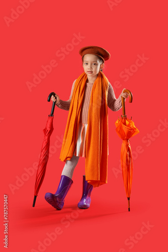 Little Asian girl in rubber boots with umbrellas on red background
