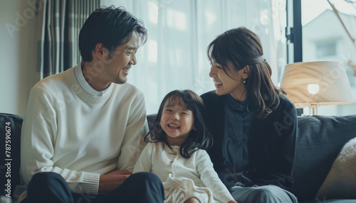 A young Japanese couple and their daughter, all smiling at the camera. Loving mother and father with kids enjoying time together at home sofa