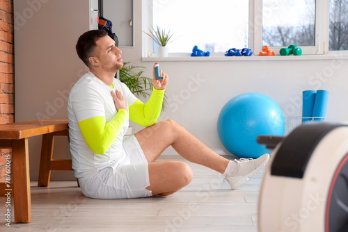 Sporty young man with inhaler sitting on floor in gym