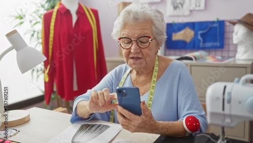 A senior woman tailor surprises at her smartphone in a colorful sewing studio surrounded by fashion designs and a sewing machine. photo