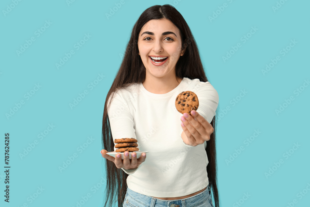 Beautiful young happy woman holding plate of tasty cookies with chocolate chips on blue background Beautiful young happy woman holding plate of tasty cookies with chocolate chips on blue background