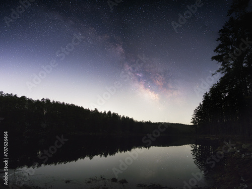 The Milky Way Galaxy arching over a lake in a forest. Stars are reflected in the water. The core of the galaxy and stars can be seen in the sky. Rocks and grassed surround the lake as well as trees.