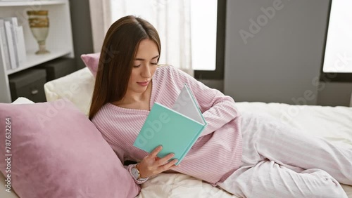 A woman in pink pajamas enjoys reading a turquoise book while reclining on a bed with pink pillows in a well-lit bedroom photo