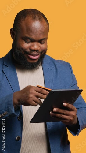 Vertical video Irked man upset after lacking money while trying to buy stuff online. Grumpy person looking at debit card after getting transaction rejected on tablet, doing facepalm, studio background photo