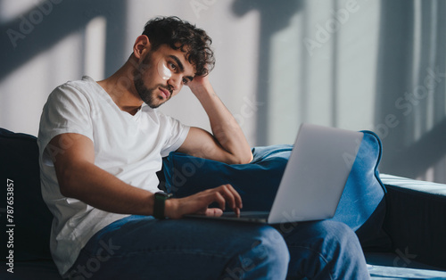 Cheerful businessman working on laptop oline looking at laptop screen sitting on sofa at home. Freelancer man working on computer distantly indoors. Freelance photo
