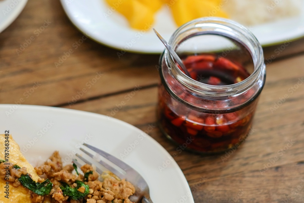 A plate of pad kra pao (holy basil pork mince stir-fry) with omelette and mixed grain rice, a jar of chilli oil, and fresh mango slices with sticky rice at a street food stall in Bangkok, Thailand