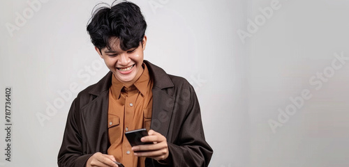 A cheerful young man in a brown shirt and a sleek black coat, his black hair styled beautifully, chuckling while browsing through his phone against a white background photo