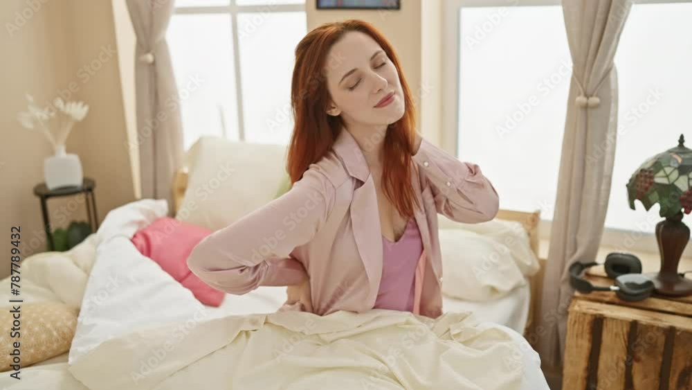 A young redhead woman experiences back pain while sitting on her bed in a bedroom. A young redhead woman experiences back pain while sitting on her bed in a bedroom.