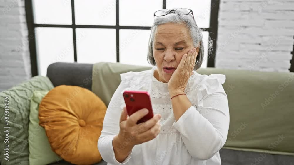 A mature woman reacts with surprise while looking at her smartphone in her cozy living room. A mature woman reacts with surprise while looking at her smartphone in her cozy living room.