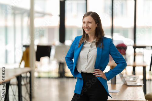 Young businesswoman with a confident smile standing with hands on hips, looking away in a modern brightly lit office environment, projecting professionalism and success Free Download Young businesswoman with a confident smile standing with hands on hips, looking away in a modern brightly lit office environment, projecting professionalism and success - GFXLoad